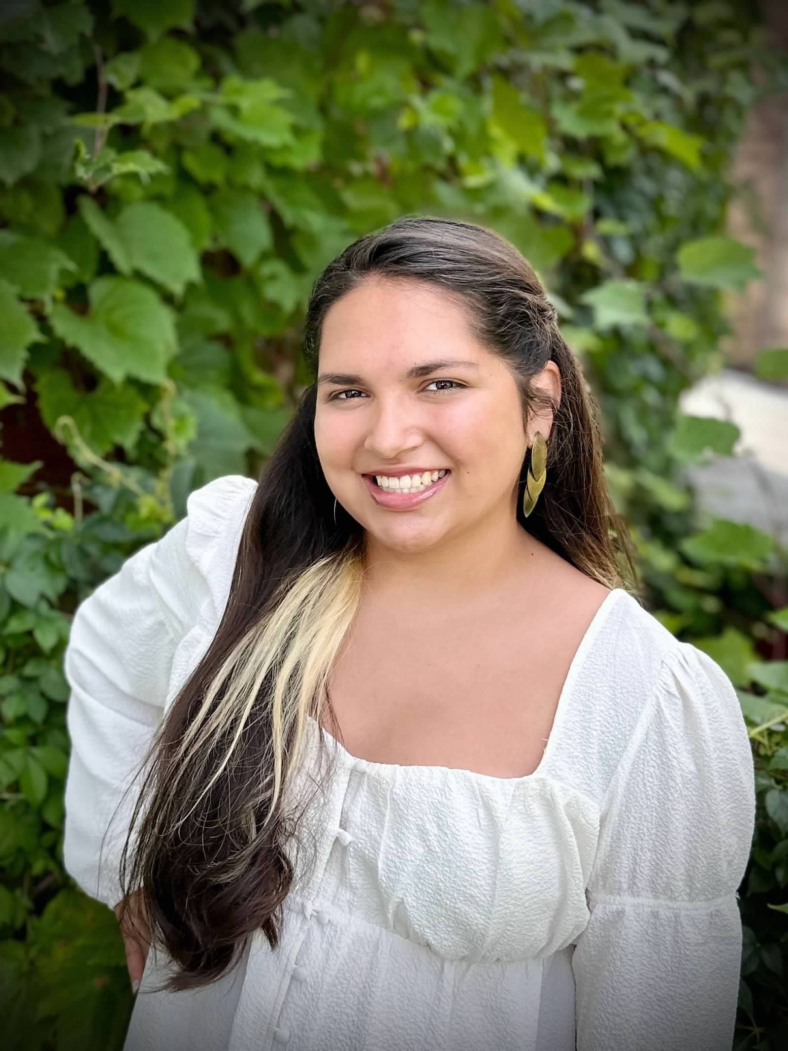 Woman with long brown hair and white dress smiling at the camera with greenery in the background